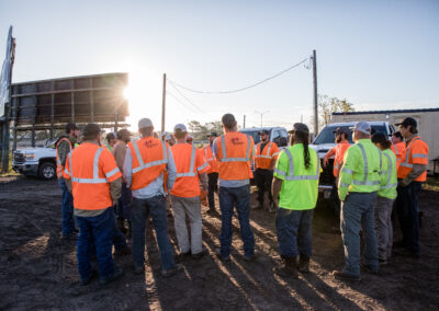 Group of men in orange and yellow safety vests with backs facing the camera as sun sets.