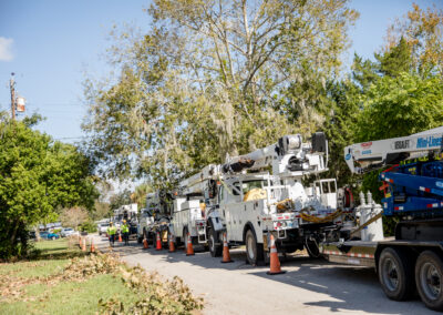 Line of bucket trucks on a narrow road with orange safety cones.