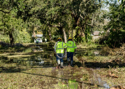 Two men in yellow safety vests walking through flooded grass.